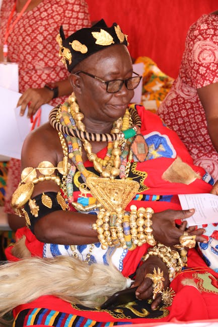 A woman dressed in traditional Maasai attire, seated outdoors during a cultural ceremony or gathering. She is adorned with elaborate jewelry, including multiple strands of beads, large gold-colored ne